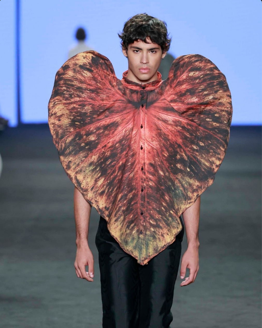 Pessoa desfila em uma passarela com uma peça artística em forma de folha, de aparência orgânica e cores terrosas, contrastando com uma calça preta e fundo desfocado.