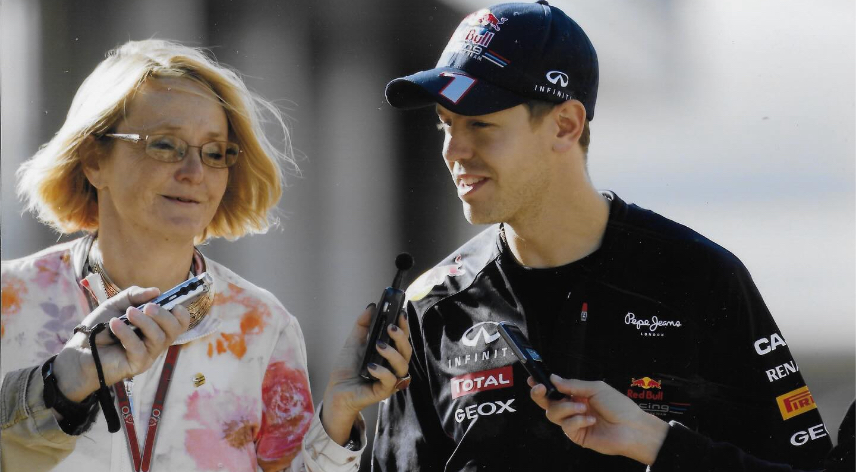 Duas pessoas adultas em ambiente externo durante uma entrevista. À direita, Sebastian Vettel veste boné e jaqueta preta com logotipos de uma equipe de Fórmula 1 e fala enquanto segura um microfone. À esquerda, a jornalista Karin Sturm segura um gravador portátil. Um terceiro microfone aparece apontado para quem responde. O fundo está desfocado.