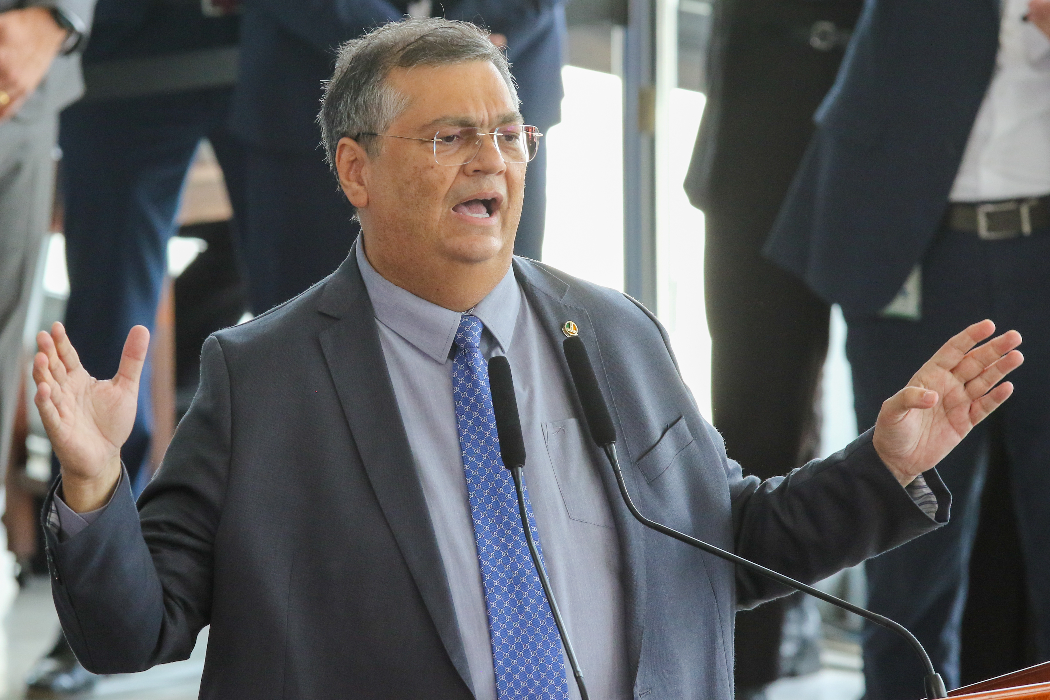 Flavio Dino discursa ao microfone, com as mãos abertas, em ambiente institucional