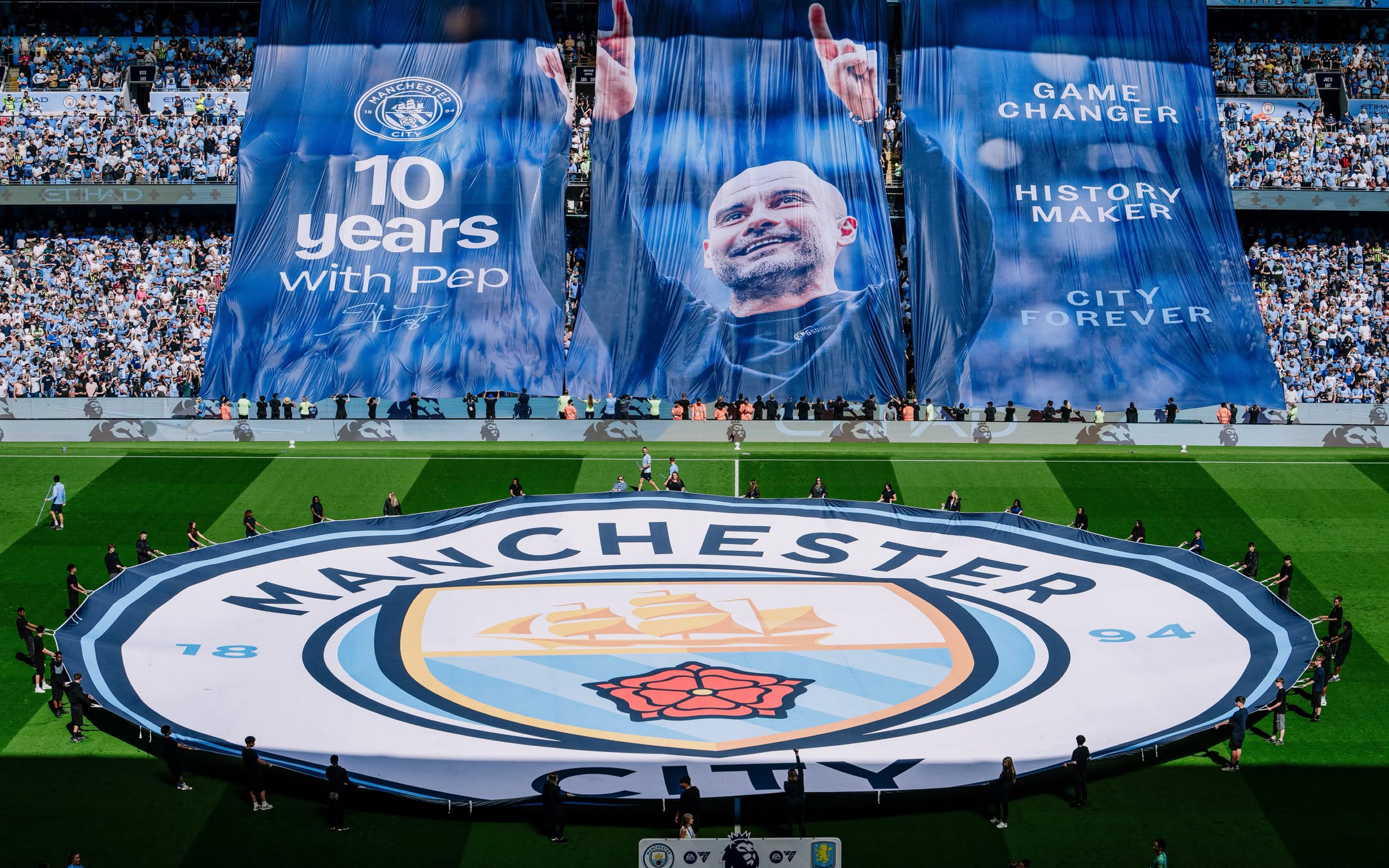 A imagem mostra o campo do Etihad com uma bandeira do escudo do Manchester City. Na arquibancada tem três bandeiras que formam uma imagem com Guardiola e os dizeres 10 years with pap