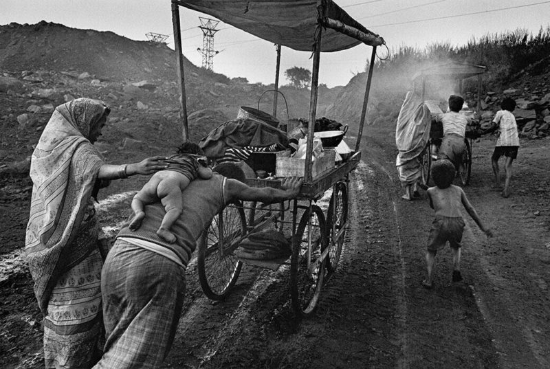 Trabalhadores de empurrando uma carroça, um deles com um bebê na costa. Alguns andando ao lado. Trabalhadores da industria de carvão na India. Foto em preto e branco