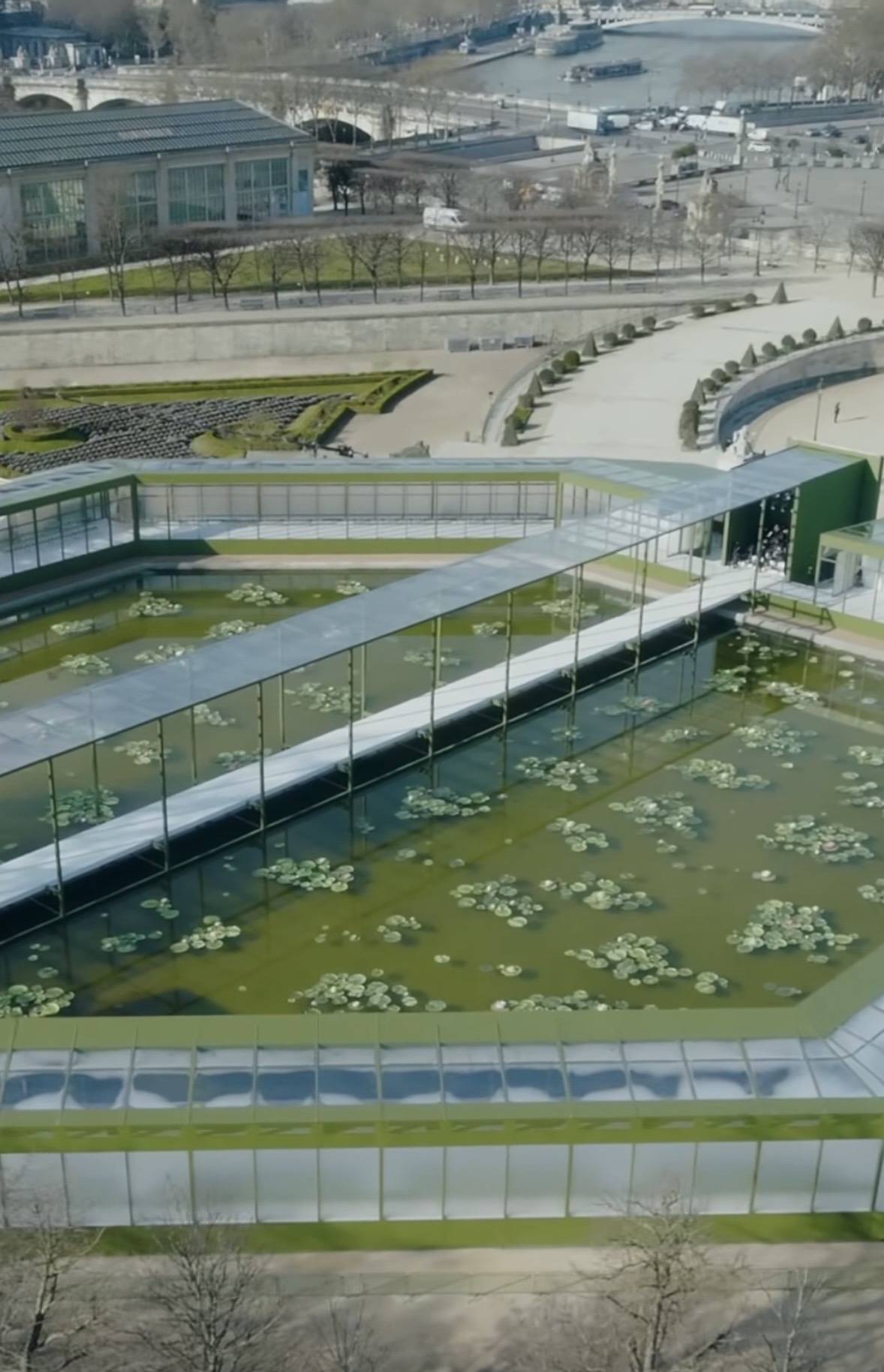 Vista aérea do palco da Paris Fashion Week 2026, com passarelas de vidro sobre espelhos d’água esverdeados, cercados por estruturas modernas e plantas aquáticas.