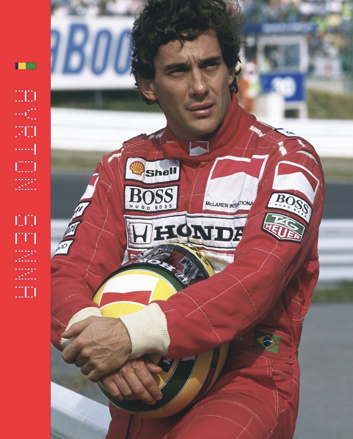 Ayrton Senna está sentado à beira da pista, vestindo um macacão vermelho da McLaren com logotipos de patrocinadores. Ele segura um capacete com as cores da bandeira do Brasil e olha para o lado com expressão séria. Ao fundo, o circuito aparece desfocado, e à esquerda há uma faixa vermelha com o nome “Ayrton Senna”.