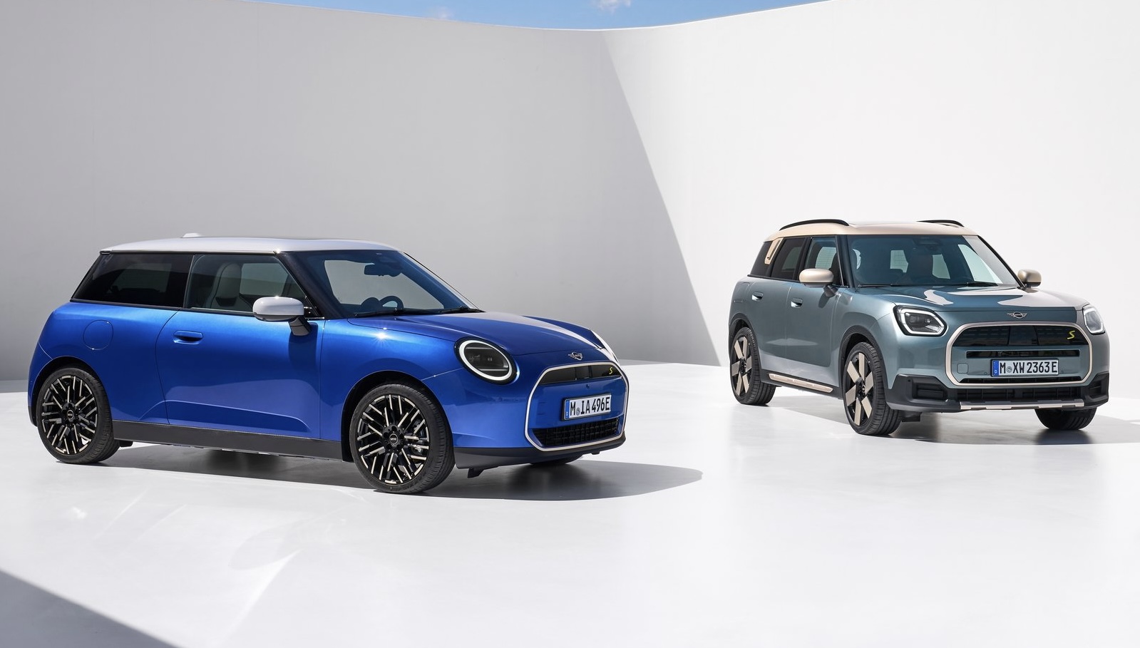 Novo Mini Cooper elétrico de lado na cor azul e teto branco a frente, e novo Mini Countryman ao fundo na cor cinza com teto dourado, ambos em um cenário totalmente branco