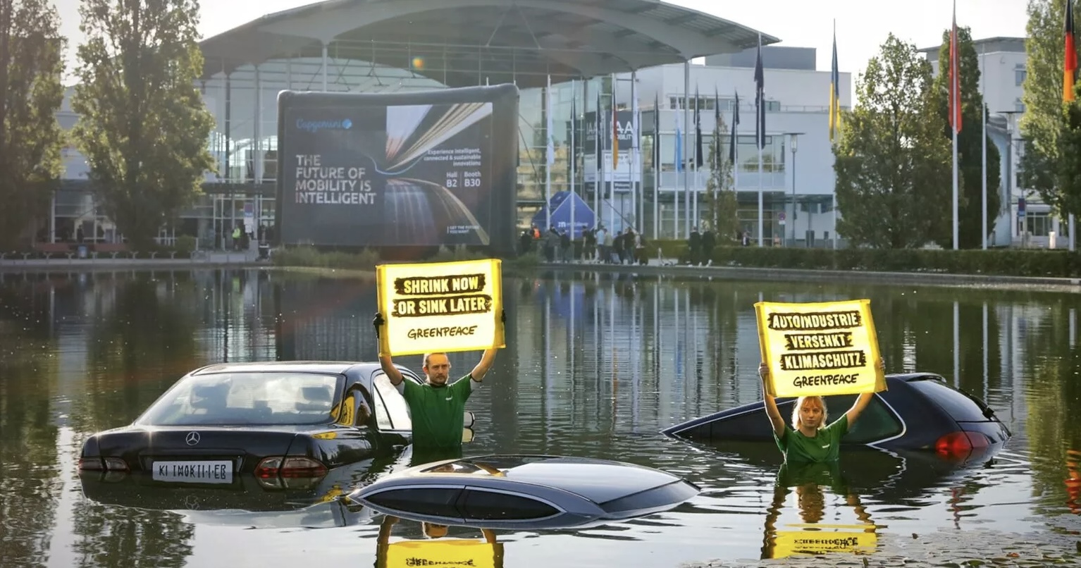 Duas pessoas segurando cartazes com escritos em Alemão, dentro de um lago com três carros parcialmente submersos, ao fundo o centro de exposições do Salão de Munique 2023 com uma fila de bandeiras de vários países ate a entrada, a qual conta com uma placa divulgando o evento escrita em inglês.