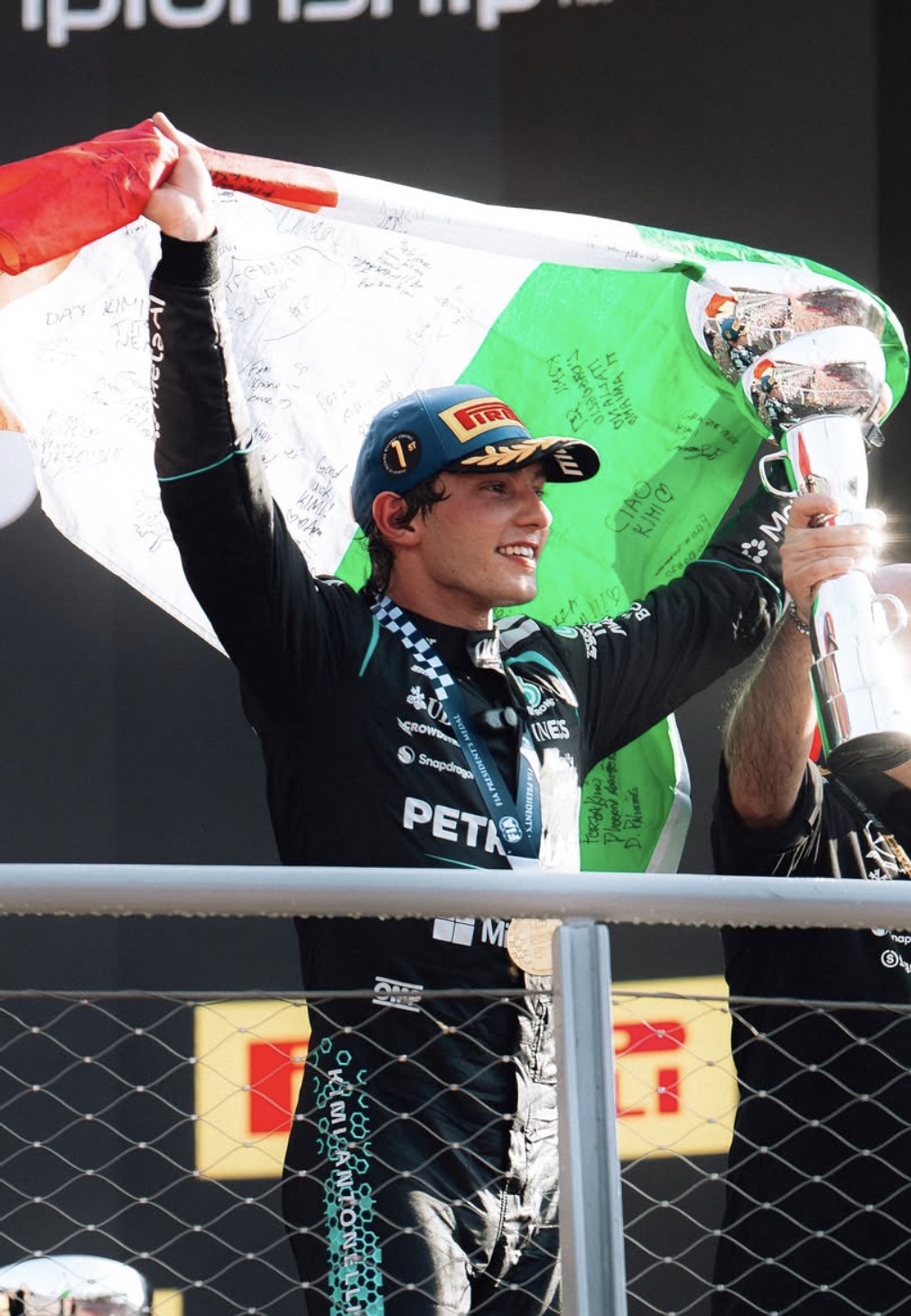 Kimi Antonelli, usando o macacão preto da Mercedes, sorri enquanto ergue o troféu e uma bandeira italiana assinada durante a celebração de sua vitória no GP de Monza.