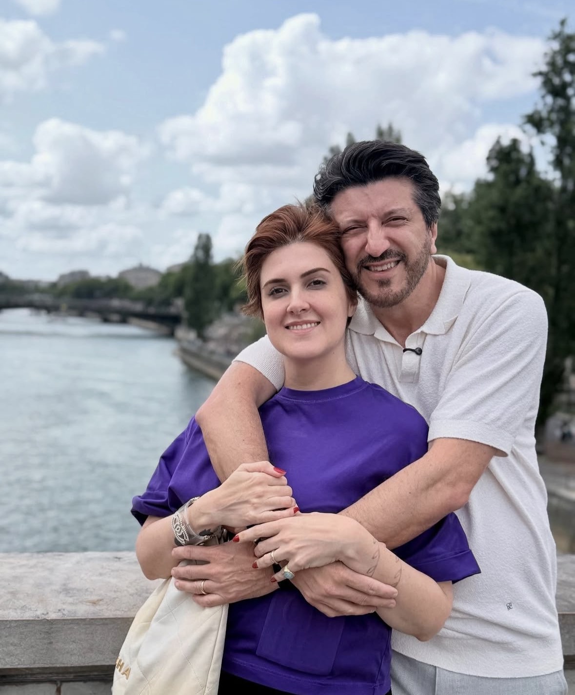 Lito Sousa e Mila Seidl posam abraçados em uma ponte sobre um rio. Ao fundo, vê-se a paisagem urbana com árvores, a água e um céu nublado, em uma cena de viagem ao ar livre.