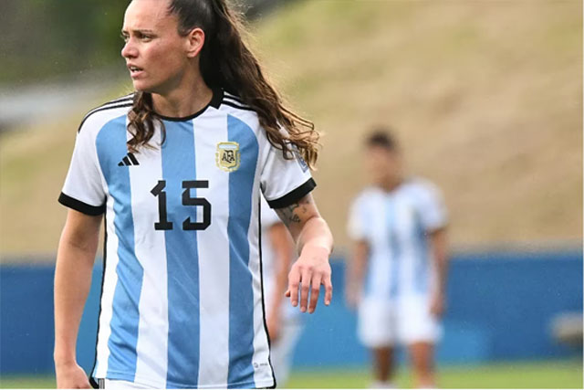 Jogadora loira, camisa 15, em campo com grama verde e natural. 