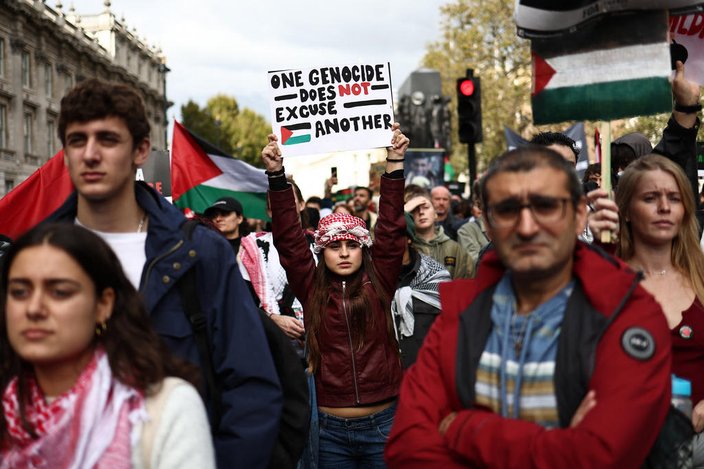 Multidão de pessoas brancas em manifestação em uma rua de Londres com um semáforo ao fundo, segurando bandeiras da Palestina com uma mulher em foco de jaqueta vermelha e bandana branca e vermelha com os braços para o alto segurando um cartaz escrito: um genocídio não justifica o outro, em inglês.