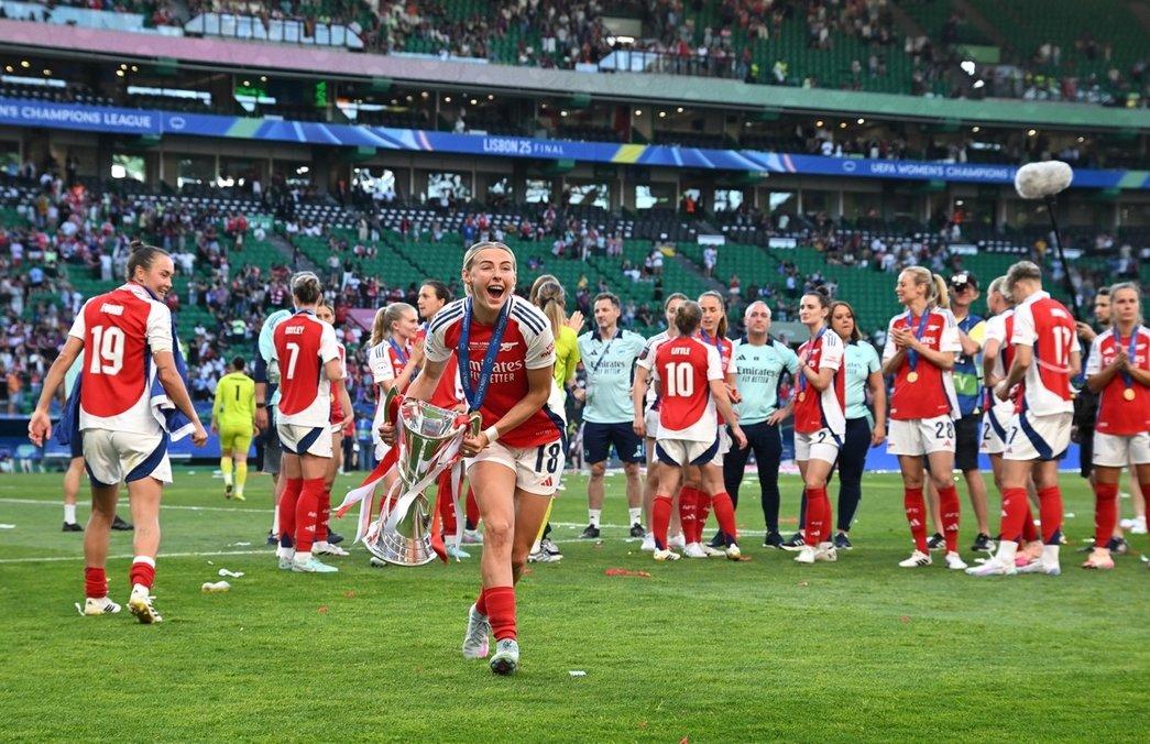 A imagem mostra a jogadota Chloe Kelly caminhando e sorrindo no campo com a medalha e a taça da Liga dos Campeões Feminina. Ao fundo estão outras jogadoras e menbros da comissão técnica do Arsenal interagindo. Mais ao fundo é possível há parte da arquibancada do estádio.