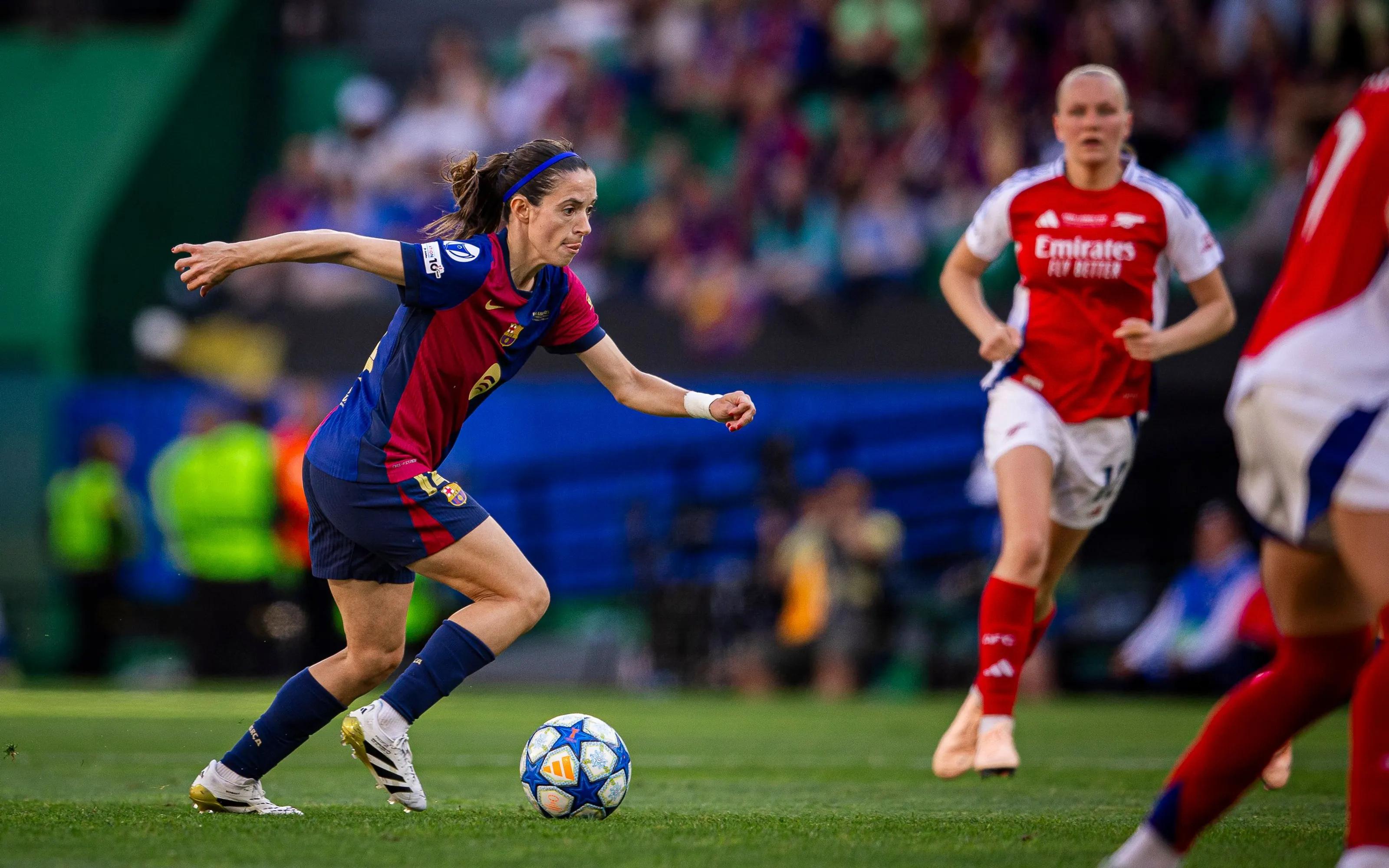 A imagem mostra a jogadora Aitana Bonmatí conduzindo a bola para a direita. Ao fundo, do lado direito, está a jogadora Maanum olhando para a jogadora do Barcelona.