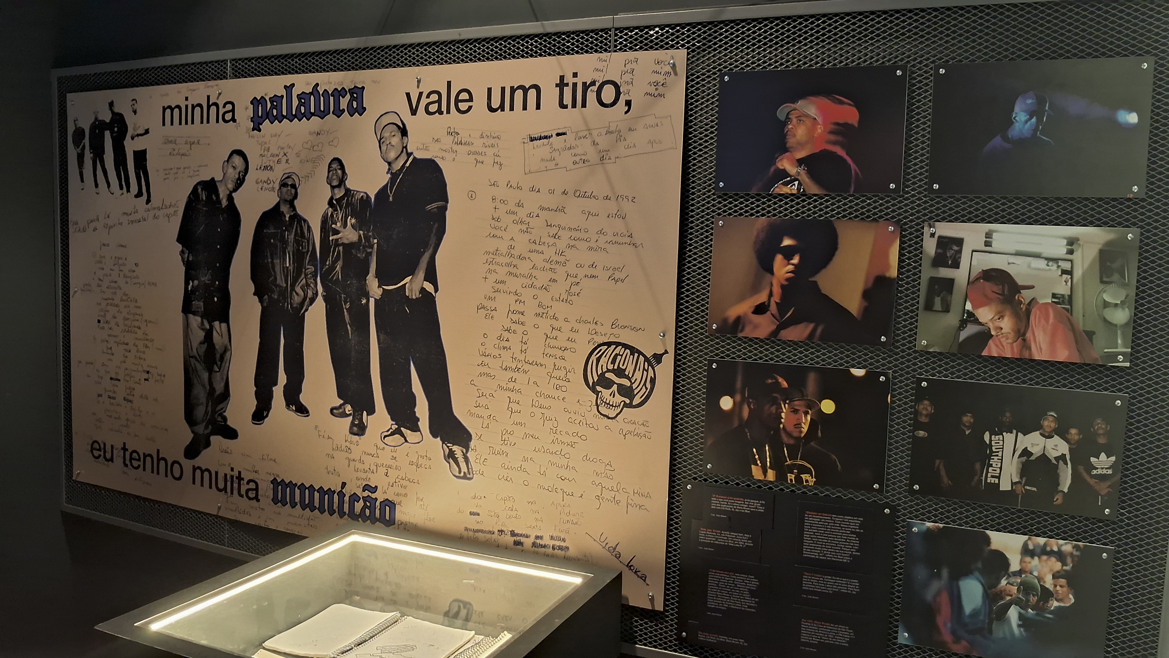 A imagem mostra um painel com os dizeres "Minha palavra vale um tiro, eu tenho muita munição" e uma foto dos quatro membros dos Racionais MC's. Ao lado, há fotos individuais dos membros.