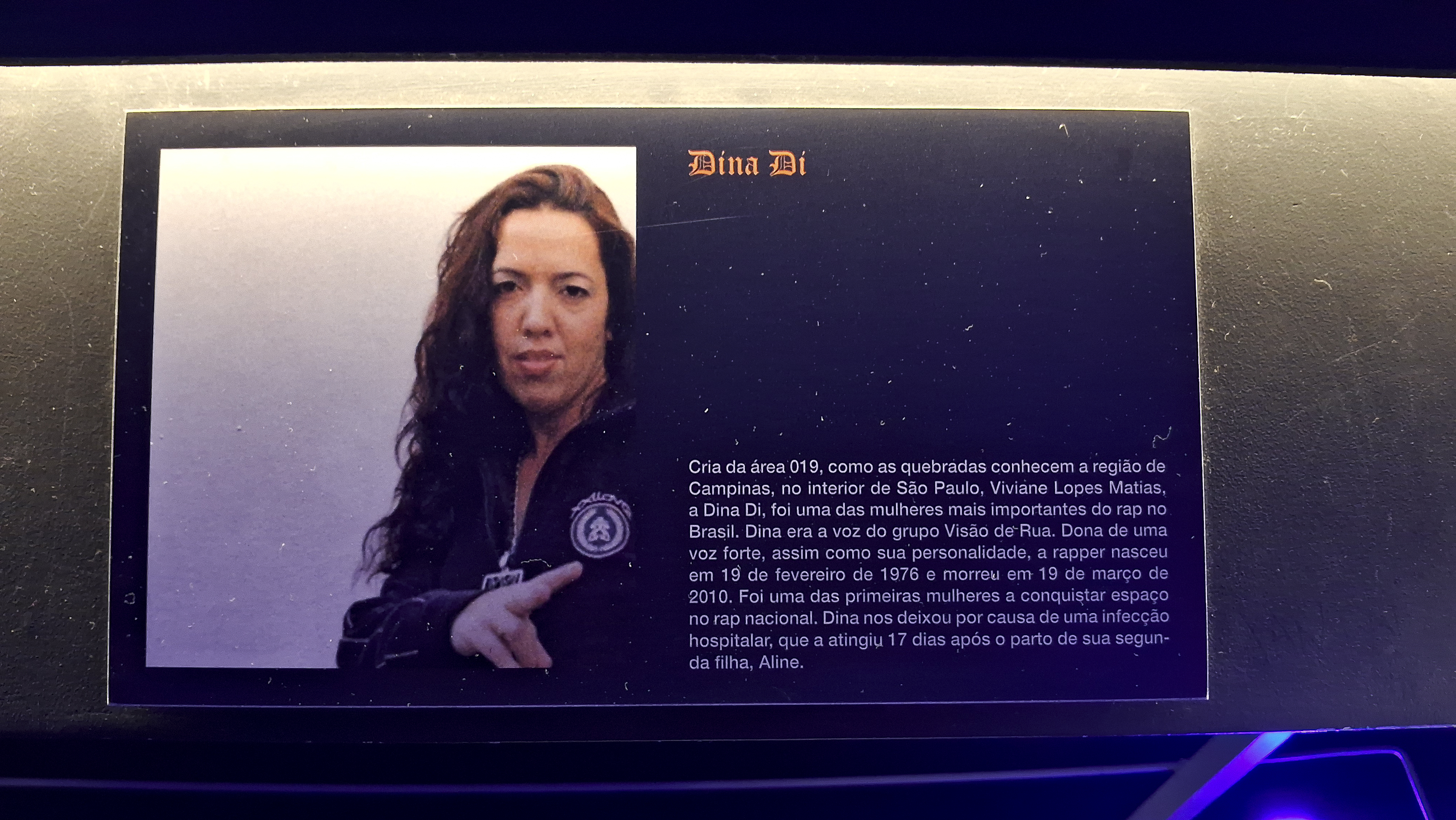 A imagem mostra uma pequena placa com a foto da Dina Di e os dizeres: "Dina Di. Cria da área 019, como as quebradas conhecem a região de Campinas, no interior de São Paulo, Viviane Lopes Matias, a Dina Di, foi uma das mulheres mais importantes do rap no Brasil. Dina era a voz do grupo Visão de Rua. Dona de uma voz forte, assim como sua personalidade, a rapper nasceu em 19 de fevereiro de 1976 e morreu em 19 de março de 2010. Foi uma das primeiras mulheres a conquistar espaço no rap nacional. Dina nos deixou por causa de uma infecção hospitalar, que a atingiu 17 dias após o parto de sua segunda filha, Aline."