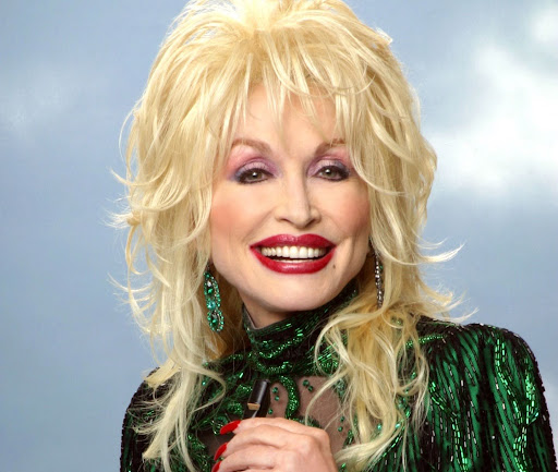 Cantora country Dolly Parton sorrindo em frente um fundo azul, usando roupas verdes e unhas pintadas de vermelho