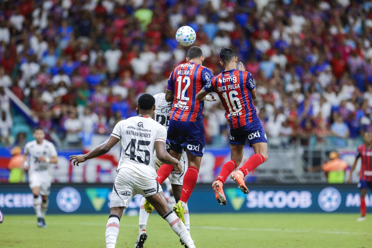 A imagem mostra os jogadores Erick Pulga e Willian José, ambos do Bahia, pulando para cabecear a bola. Logo atrás está o jogador Nathan Mendes, do Vitória, olhando para os jogadores. Ao fundo está a torcida.
