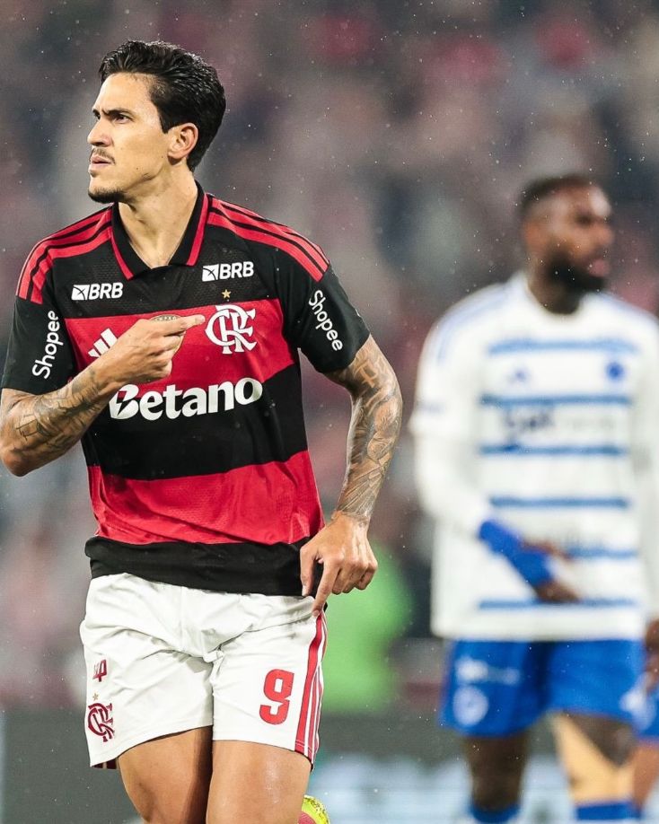A imagem mostra à esquerda o jogador Pedro, do Flamengo, apontando com o dedo para o escudo do time em sua camisa. Na direita ao fundo, está o jogador Gerson, do Cruzeiro, com efeito de edição de desfoque.