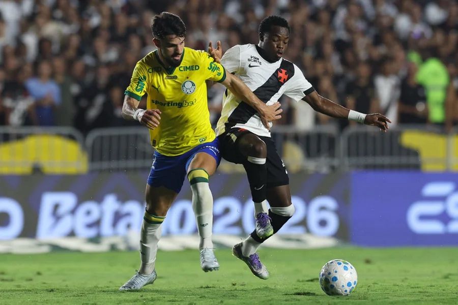 A imagem mostra o jogador Flaco López, do Palmeiras, e o jogador Andrés Gómez, do Vasco, disputando a bola. Ao fundo está a torcida.