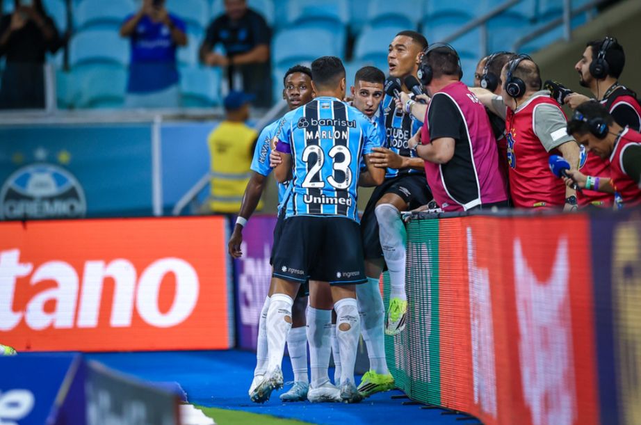 A imagem mostra jogadores do Grêmio comemorando o gol nas placas de publicidade. Na direita dos jogadores estão alguns jornalistas.