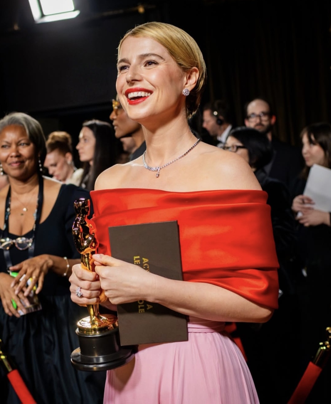 Jessie Buckley sorri enquanto segura uma estatueta do Oscar em um evento formal. Ela usa um vestido elegante com parte superior vermelha tomara que caia e saia rosa clara, além de brincos e colar delicados. Ao fundo, há outras pessoas bem vestidas e luzes de bastidores, criando um clima de premiação e celebração.