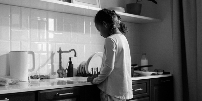 A imagem, em preto e branco, mostra uma menina de costas olhando para uma pia.