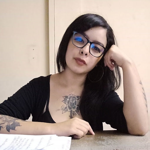 Mulher branca de cabelos pretos um pouco abaixo da linha do ombro . Está usando uma blusa preta, óculos pretos, um brinco de argola na orelha esquerda, e um piercing na região do nariz. Possui uma tatuagem de flor na região do peito, outra no braço direito