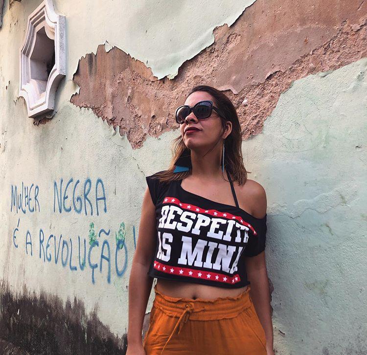 Paloma Queiroz está usando uma camisa que traz a frase: "respeita as mina" e atrás dela uma parede grafitada com a frase: "mulher negra é a revolução"