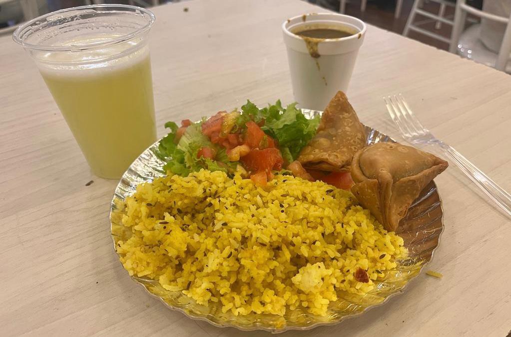 Prato de papel alumínio com arroz temperado amarelo, samosas de legumes e uma salada de alface crespa e tomates em cubo, sob uma mesa bege de madeira junto a um garfo de plástico transparente, copo de papel com sopa de lentilhas, e um copo de plástico transparente com o suco Nimbu Pani indiano de aparência verde 