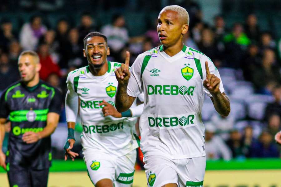 Homem comemora gol, usando uniforme branco e verde, com dedos para cima.