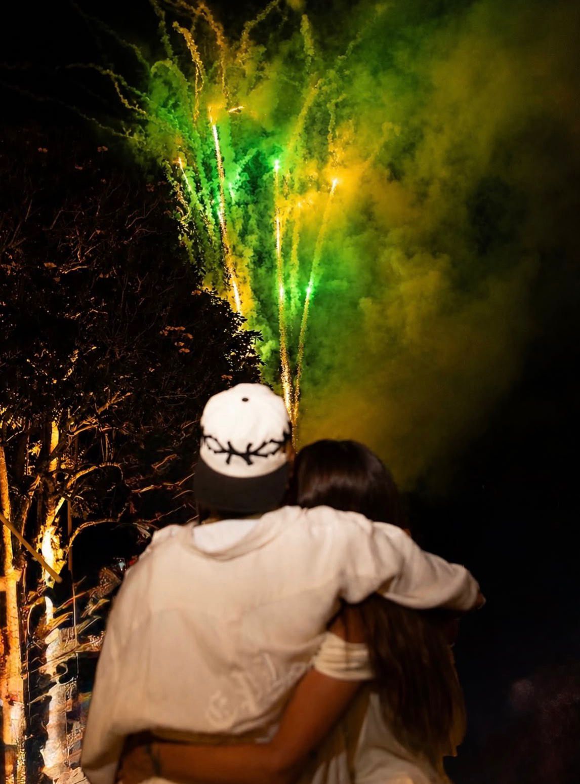 Neymar abraçado com a esposa, Bruna Biancardi, de costas para a câmera. Explosões de fogos nas cores verde e amarelo são destaque ao fundo.